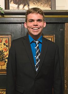A young person in a suit smiling is standing in front of a wall covered with art in frames